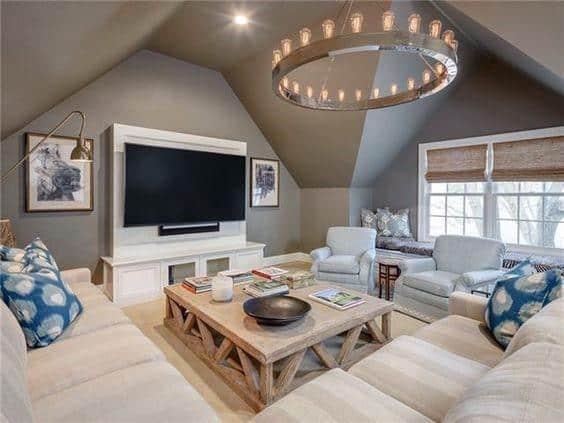 Cozy attic living space with sectional sofa, wooden coffee table, chandelier, and wall-mounted TV.