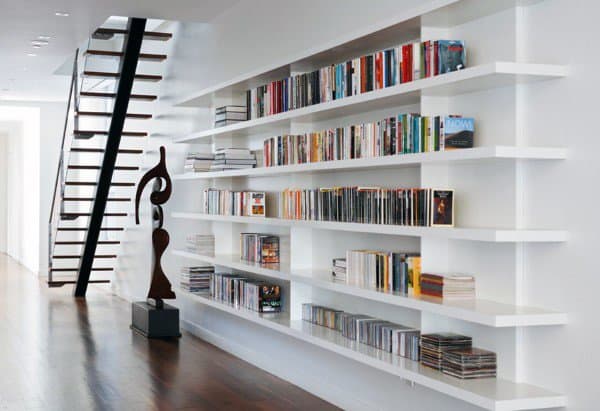 Modern interior with white shelves full of books and CDs, a wooden staircase, and abstract sculpture on wooden floor
