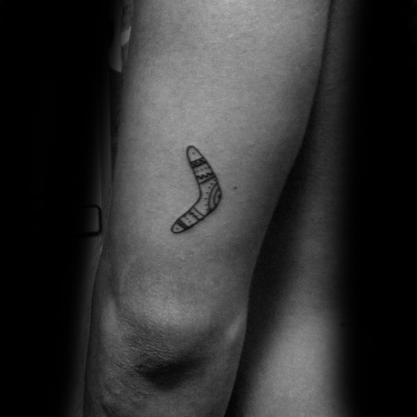 Small minimalist boomerang tattoo with fine line tribal-style patterns, placed on the back of the upper arm.
