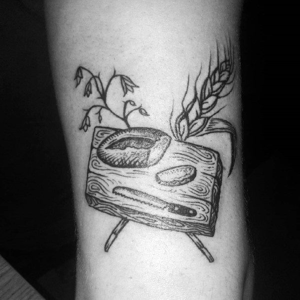 Black ink tattoo of a rustic wooden cutting board with a loaf of bread, a knife, wheat, and plants, creating a vintage farmhouse aesthetic.
