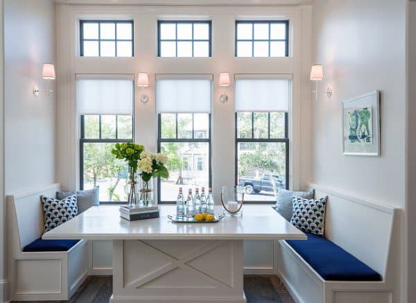 Cozy breakfast nook featuring a white table, blue benches, and large windows overlooking lush greenery