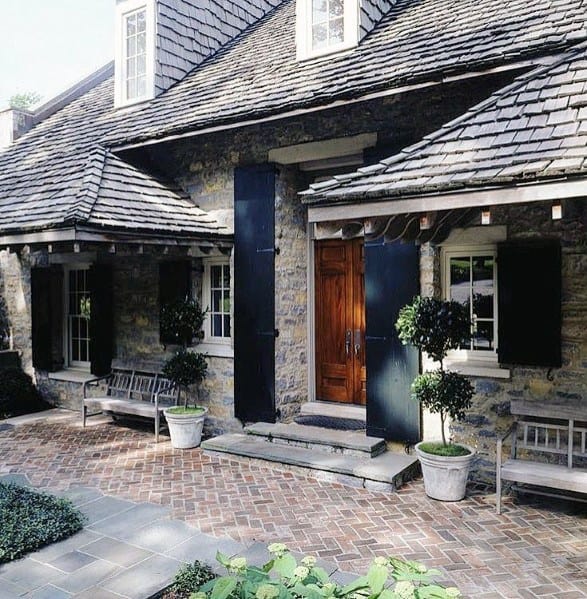 Elegant brick walkway leading to a stone home with dark shutters, planters, and benches for a welcoming entrance