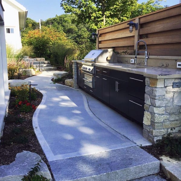 Outdoor kitchen with built-in grill and sink, stone and wood accents, set along a curved concrete pathway, lush greenery surrounds