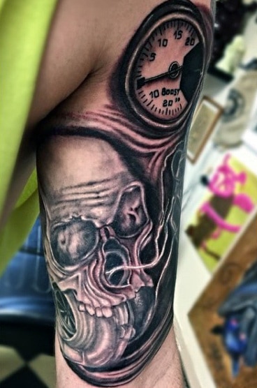 Tattoo of a skull and pressure gauge on an upper arm with a black and grey color scheme