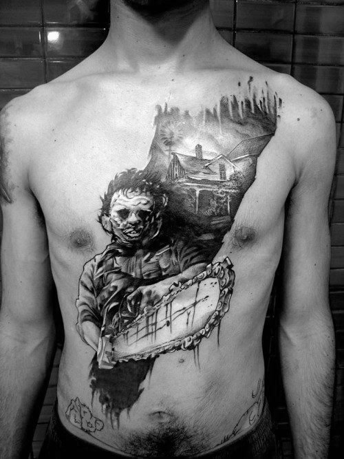 Realistic black and gray chest tattoo of Leatherface holding a chainsaw, with a haunted house and forest silhouette in the background.