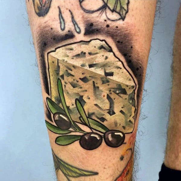 Colorful tattoo of a wedge of feta cheese with black olives and olive leaves, featuring bold outlines, rich shading, and a textured background.