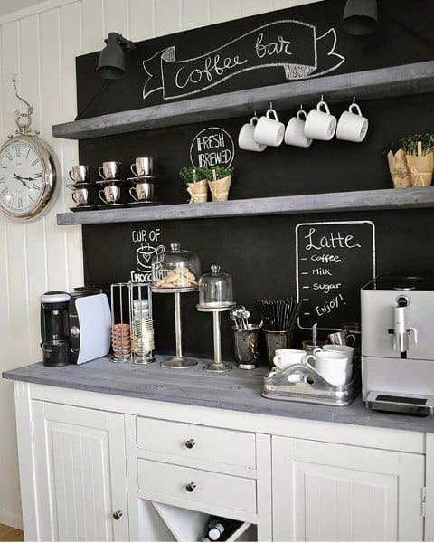 White coffee bar with chalkboard wall, wooden shelves, mugs, and coffee machines.