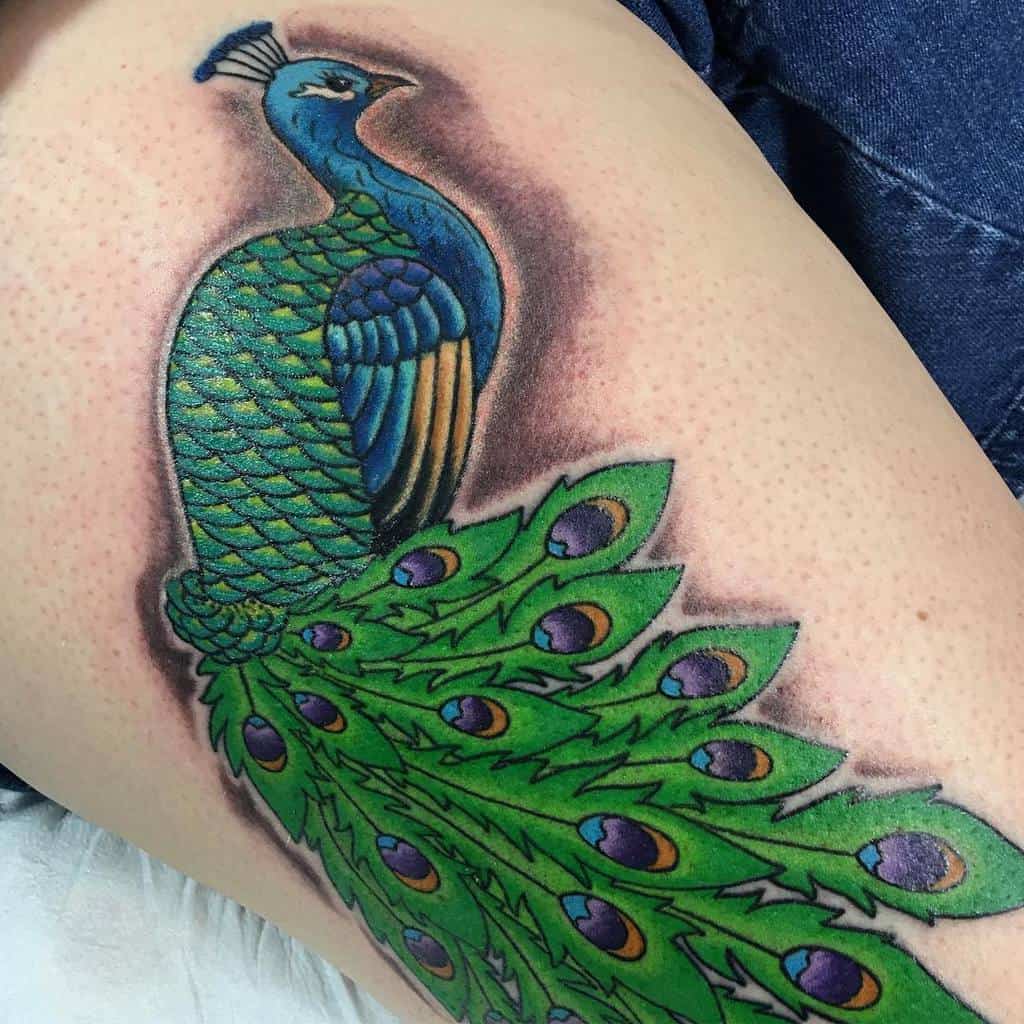 Vibrant peacock tattoo on the thigh with detailed green and blue feathers, showcasing a bold and colorful design with intricate eye patterns in the tail feathers.