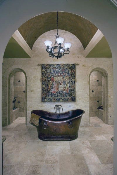 Elegant bathroom with a bronze tub, shower, ornate chandelier, tapestry on stone wall, and arched doorways