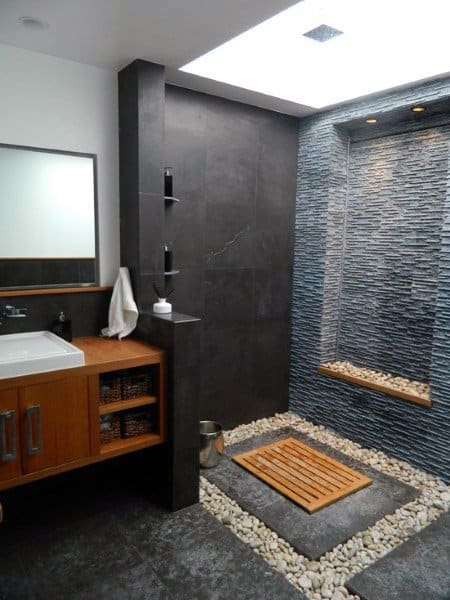 Modern bathroom with a sleek shower, dark stone wall, wooden floor mat, pebble border, and a stylish vanity