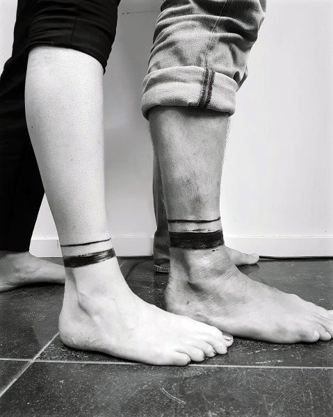 Matching ankle tattoos of two bold black bands, symbolizing strength, unity, and a minimalist expression of commitment.