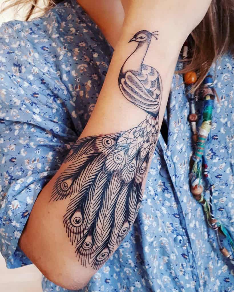 Detailed black and white peacock tattoo with flowing feathers and intricate eye patterns, covering the forearm.