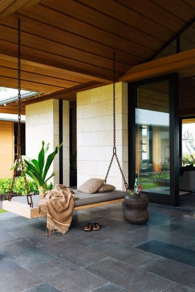 Outdoor hanging daybed with chains, cozy cushions, and greenery under a wooden patio roof.