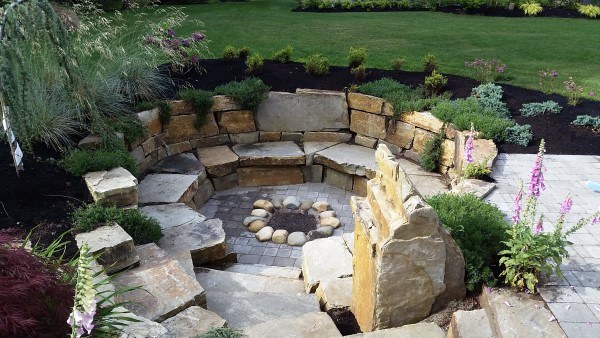 Fire pit surrounded by stone seating in a landscaped garden, bordered by plants and a grassy lawn
