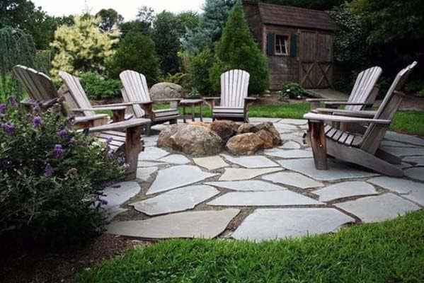 Wooden chairs around a stone fire pit in a garden with lush greenery and a wooden shed in the background
