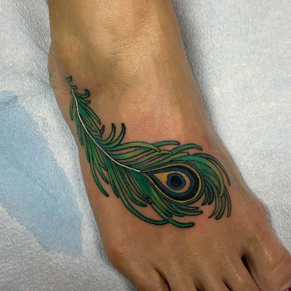 Colorful peacock feather tattoo on the foot, featuring rich green, blue, and gold hues with bold outlines and intricate details.