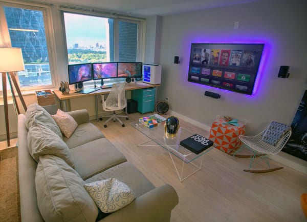 A modern home office with dual monitors, a light gray sofa, a wall-mounted TV, and colorful decor