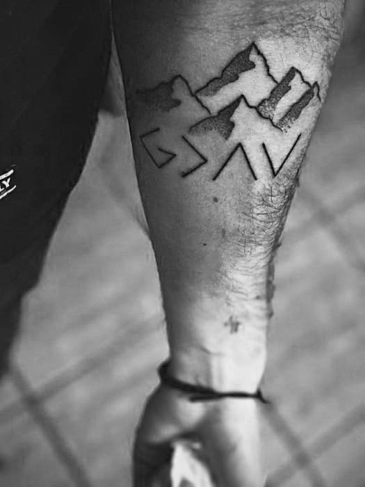 Forearm tattoo featuring "God is greater than the highs and lows" symbol with detailed mountain silhouettes