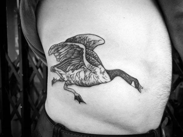 Black and white tattoo of a goose in mid-flight, inked on a person’s ribcage, with detailed wings and shading for a realistic look.