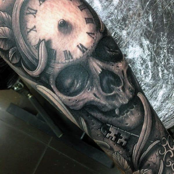 Tattoo of skull with Roman numeral clock on forehead and cross necklace on an arm