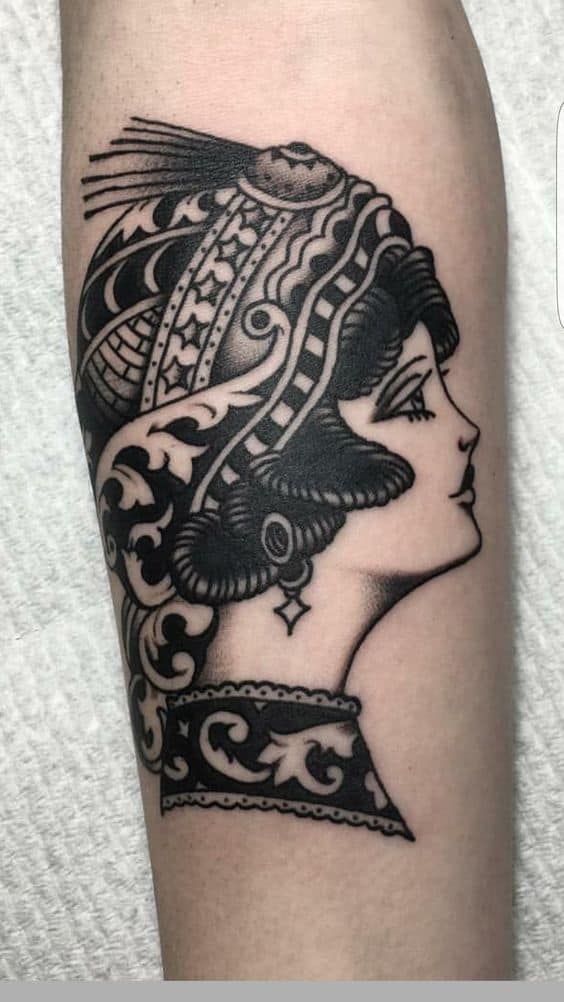 Ornate black and grey tattoo of a gypsy woman in profile with an intricate headpiece, bold shading, and elegant patterns on the forearm.