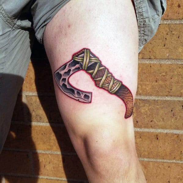 Curved hatchet tattoo on the thigh with a rugged, hammered metal blade and a wrapped handle, designed with bold shading and red accents.