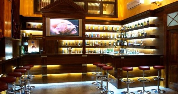 A cozy bar with wooden decor, a row of stools, shelves lined with bottles, and a TV displaying an image above