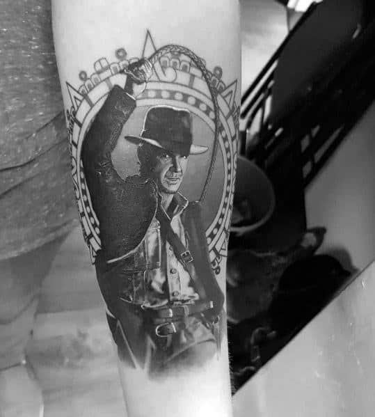 Black and gray Indiana Jones tattoo showing him mid-action with a whip, framed by an intricate circular design.