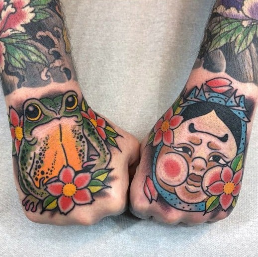 Traditional Japanese-style hand tattoos featuring a vibrant orange frog on one hand and a smiling Daruma-like face on the other, adorned with flowers.