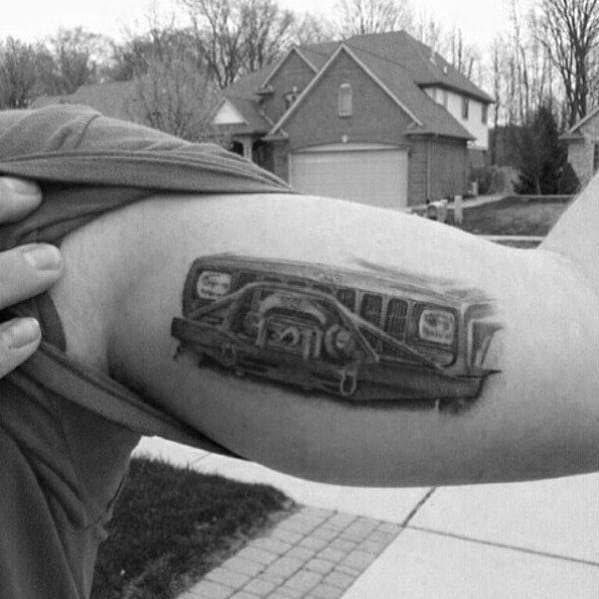 Black and grey Jeep tattoo on the bicep, featuring a detailed front grille and winch, creating a bold and rugged off-road design.