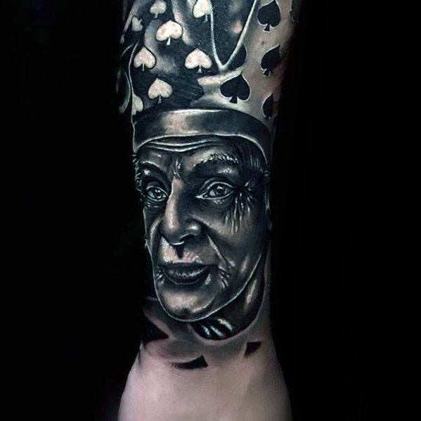 A black and gray tattoo of a jester's face, wearing a crown adorned with spade symbols, capturing a detailed and expressive portrait.