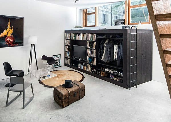 Compact studio with loft bed, built-in shelves, ladder, and vintage trunk as a coffee table.