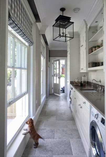 Bright laundry room with large windows, white cabinets, stone flooring, and a small dog by the window.