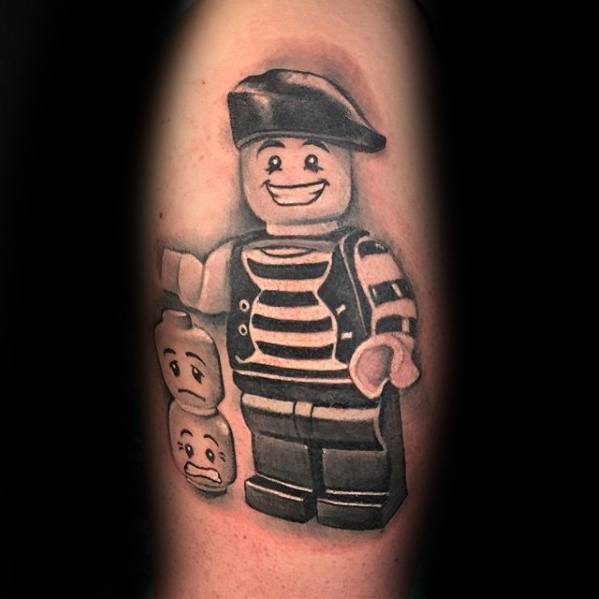 Black and grey tattoo of a smiling Lego mime figure with striped shirt, beret, and two interchangeable heads showing sad and surprised faces.