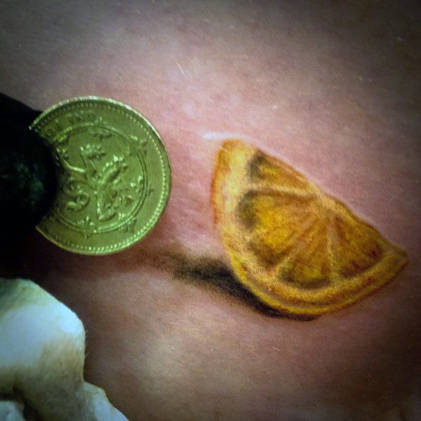 Realistic tattoo of a tiny lemon wedge with soft shading, placed beside a coin for scale to show its miniature size.