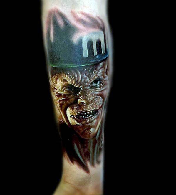 Realistic horror-style leprechaun tattoo with snarling face, wrinkled skin, and a dark hat marked with a bold white “M”.