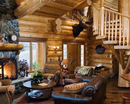 Rustic log cabin living room with stone fireplace, wooden staircase, and leather furniture.