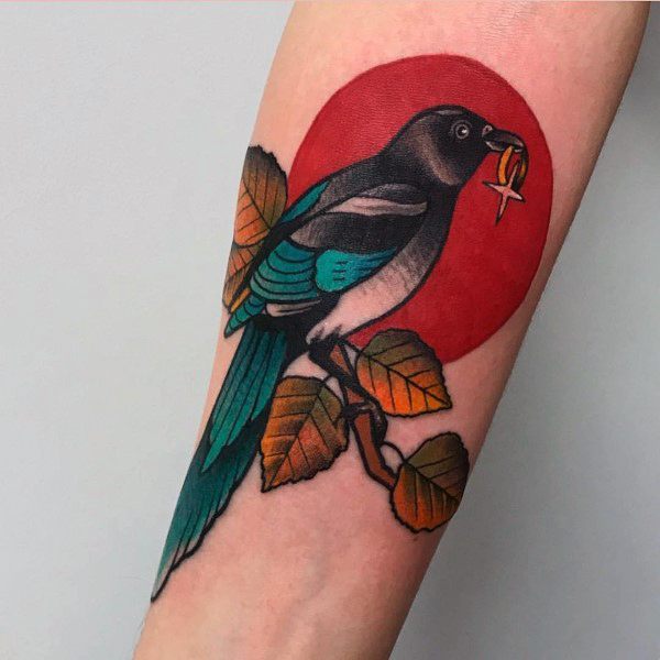 Magpie tattoo on forearm with a red circle backdrop, perched on a branch with autumn leaves, holding a gold ring in its beak.