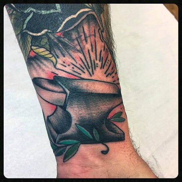Bold anvil tattoo on the wrist with black shading, green leaves, and red radiant bursts for a striking and energetic design.