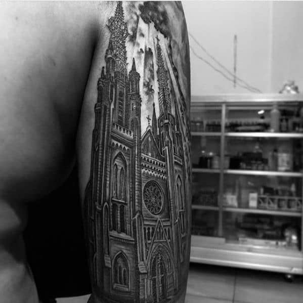 Upper arm tattoo of a grand gothic cathedral with tall spires, rose window, and dramatic clouds adding a powerful atmospheric effect.