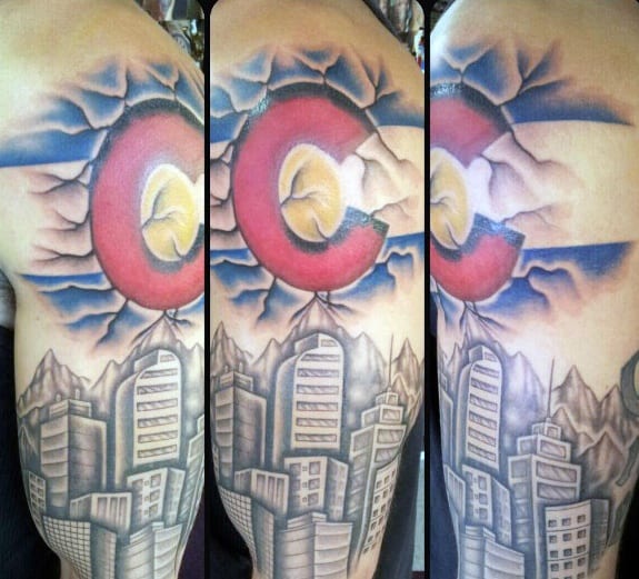 Realistic Denver skyline tattoo with mountains and a large Colorado flag breaking through the sky, wrapping around the upper arm.