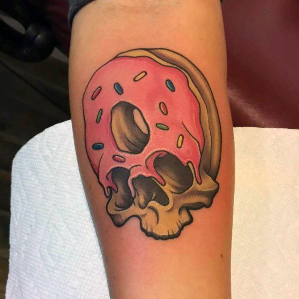 Tattoo of a pink frosted donut with colorful sprinkles melting into a detailed skull design on the forearm.