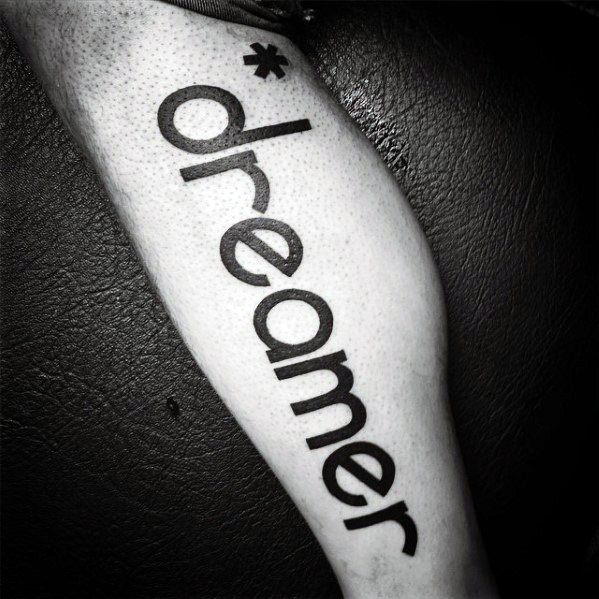 Bold lowercase tattoo of the word “*dreamer” running vertically along the leg in modern sans-serif font.