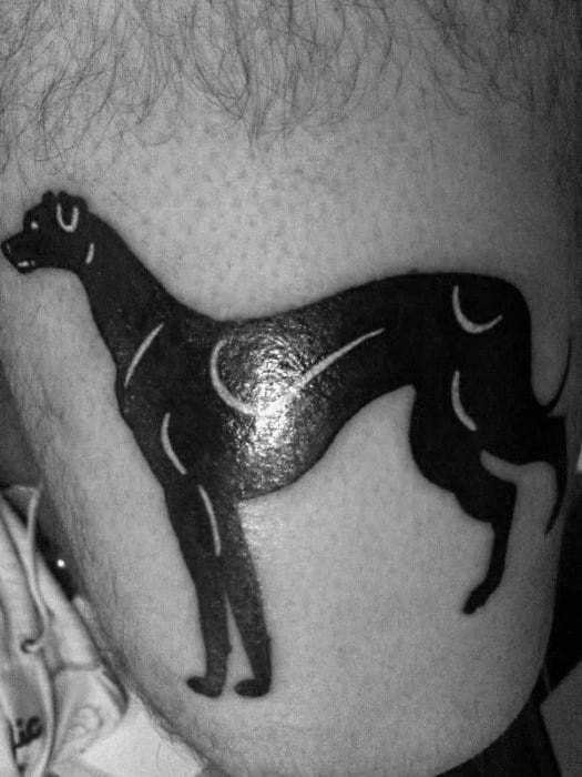 A minimalist black ink greyhound tattoo, featuring a simple, clean silhouette of the dog in profile with subtle curved details.