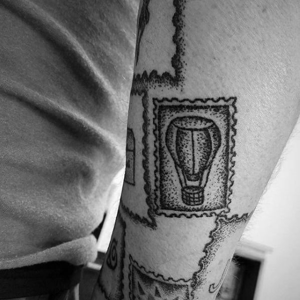 A tattoo depicting a vintage postage stamp features a classic hot air balloon design elegantly inked on the person's arm