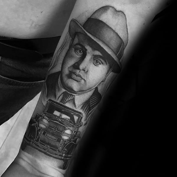 Al Capone tattoo featuring a detailed black-and-white portrait of the gangster with his signature fedora, accompanied by a vintage car beneath him.