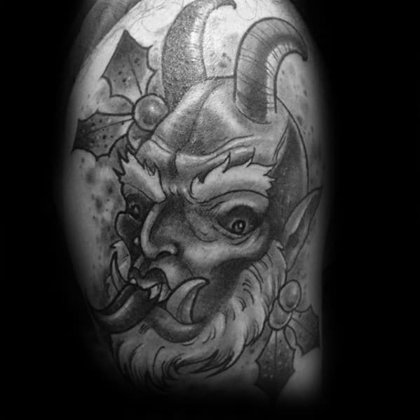 Black and grey Krampus tattoo featuring a fierce horned face with fangs, surrounded by holly leaves and a dark, moody background.