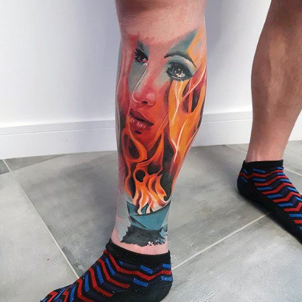 Surreal leg tattoo of a woman's face with striking eyes, blending into vivid orange flames and a fragmented landscape below.