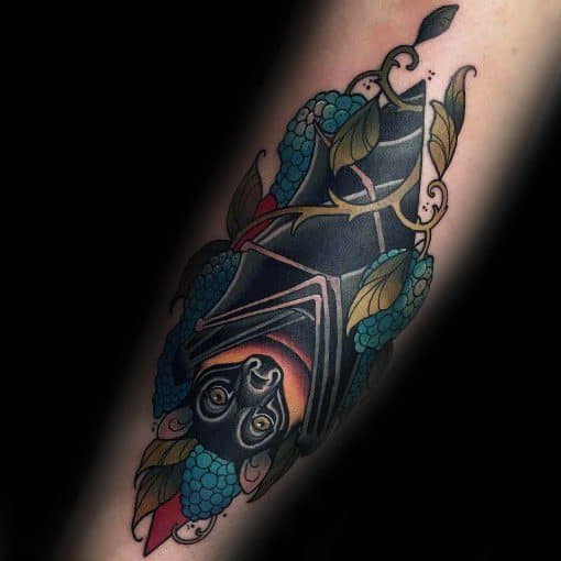 Neo-traditional tattoo of an upside-down bat wrapped in wings, surrounded by blue berries and leaves with ornate detailing.