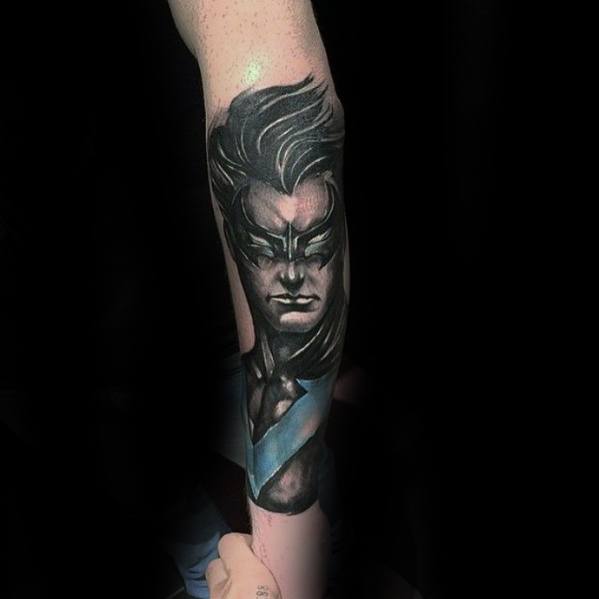 Realistic Nightwing forearm tattoo in black and gray with a bold blue chest emblem and intense facial expression.